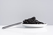 Stainless fork in full bowl of black spaghetti with cuttlefish ink on white table in studio on gray background