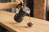 Crop anonymous male barista pouring hot aromatic Americano from stove top coffee maker into glass cup placed on wooden counter in cafe