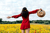 Back view anonymous trendy female in red sundress standing on blossoming field with yellow and red flowers with outstretched arms on warm summer day