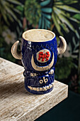 From above of bull shaped tiki mug of alcohol drink with froth placed against wooden table on blurred background