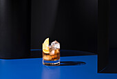 Glass of alcoholic cocktail made with red vermouth served with orange slices in light studio with blue and black wall
