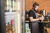 Side view of bearded male barista cleaning pitcher with napkin while standing at counter near coffee machine and working in coffee shop