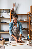 Focused adult lady in casual clothes and apron sculpting clay piece and smiling during pottery class in workshop