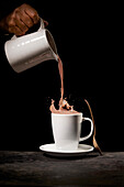 Crop unrecognizable person pouring tasty cocoa into white mug placed on table in kitchen on black background
