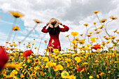 Back view anonymous trendy female in red sundress standing on blossoming field with yellow and red flowers and touching hat on warm summer day