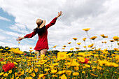 Back view anonymous trendy female in red sundress standing on blossoming field with yellow and red flowers with outstretched arms on warm summer day