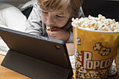 Little boy lying down comfortable on sofa with popcorn bucket while eating and watching cartoon on tablet