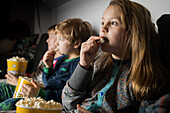 Young amazed siblings kids sitting comfortable on sofa with popcorn bucket while eating and watching TV