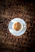 Top view of ceramic cup of coffee with foam among spilled roasted beans on wooden surface