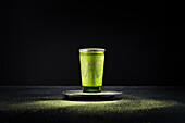 Gesunder grüner Matcha-Kräutertee, serviert in einer Glastasse mit Metallverzierung auf einer Untertasse, bestreut mit Pulver auf einem schwarzen Tisch