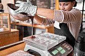 Hispanic woman in apron and gloves filling plastic mold on digital scale with liquid chocolate during work in confectionery in daytime