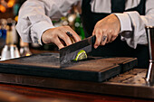 Crop young woman in uniform chopping fresh lime on counter while preparing cocktail during work in bar