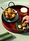 Cropped unrecognizable person hands eating traditional codfish fritters server on tray with fresh herbs and glass of beer on green background