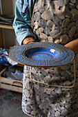 Crop unrecognizable artisan in apron demonstrating creative ceramic plate in workshop