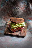 Hoher Blickwinkel auf ein appetitliches Sandwich mit frischem, knusprigem Brot über Salatblättern und Speck auf einem Holzbrett