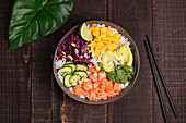 Top view of appetizing colorful poke bowl with fresh salmon and rice served with ripe avocado slices, cabbage and mango placed on wooden tray