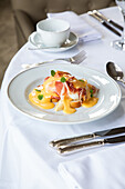 From above of appetizing Eggs Benedict dish with bacon topped with hollandaise sauce served in white plate on table with cup of coffee and silverware in restaurant during brunch