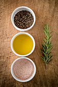 Schale mit Himalaya-Salz neben Olivenöl und gemahlenem schwarzem Pfeffer vor Rosmarinzweig auf Holzoberfläche