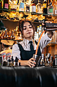 Female barkeeper pouring orange alcohol cocktail in metal shaker while preparing refreshing drink and standing at counter in bar