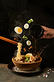 Cook holding noodles with chopsticks and adding miso broth into ramen while eggs and onion with mushrooms falling into bowl