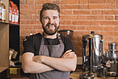 Bärtiger positiver männlicher Barista in Schürze verschränkt die Arme, während er im Kaffeehaus steht und in die Kamera schaut