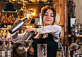 Agile professional young female bartender in uniform mixing drinks in stainless shaker while making cocktails at bar counter in restaurant