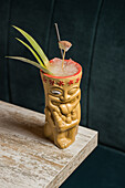 Polynesischer Tiki-Becher mit kaltem alkoholischem Getränk, dekoriert mit Strohhalm und grünen Ananasblättern, auf einem Holztisch