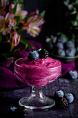 Glass with fresh blueberry mousse garnished with fresh berries served on dark table with blurred flowers