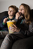Young amazed siblings kids sitting comfortable on sofa with popcorn bucket while eating and watching TV