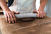 From above of crop unrecognizable male artisan in apron rolling up clay piece at table in traditional workshop