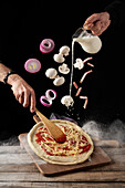 Koch mit Spachtel gießt Sahne für Soße, während geschnittene Zwiebeln und Pilze mit Speck auf Pizza mit Käse und Tomatenmark fallen
