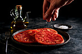 Crop anonymous person pouring sea salt on sliced fresh juicy tomato in dark bowl placed near jug of olive oil