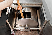 From above anonymous woman in apron using wooden stick to mix cocoa beans in drum of roasting machine during work in cafe