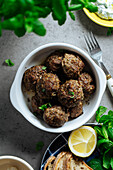 Draufsicht auf appetitliche traditionelle hausgemachte gebratene griechische Fleischbällchen, die auf grauem Hintergrund neben einem Teller mit Brot und Zitrone in der Küche serviert werden