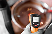 From above soft focus of anonymous worker using infrared thermometer to measure temperature of liquid chocolate in bowl in confectionery