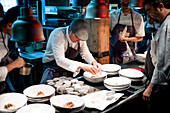 Young cooks men in aprons carefully serving meal in white ceramic dishes on restaurant kitchen