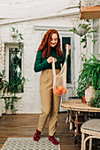 Full body of young red haired female with eco friendly shopping string bag with tangerines standing in room decorated with houseplants