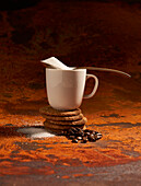 High angle of white ceramic jug with coffee and spoon full of sugar placed on pile of baked sweet oatmeal cookies near coffee beans and scattered granulated sugar