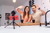 Positives Bloggerpaar, das mit der Hand in ein Handy mit Mikrofon winkt, während es zu Hause am Küchentisch mit einer Tasse Kaffee sitzt