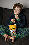Amazed little boy sitting comfortable on sofa with popcorn bucket while eating and watching TV