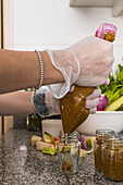 Side view of crop anonymous female in gloves pouring vegetable appetizer in glass bottle near bowl with green lettuce in kitchen at home