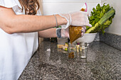 Side view of crop anonymous female in gloves pouring vegetable appetizer in glass bottle near bowl with green lettuce in kitchen at home