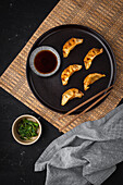 Top view of chopsticks with gyoza on plate near bowls of soy sauce and greens during lunch on black table