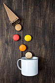 From above colorful French macaroons with various fillings falling from paper cone into a mug on wooden table
