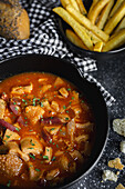 From above of tasty callos a la madrilena in black bowl near French fries and bread with seeds