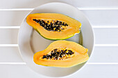 Draufsicht auf eine halbierte frische reife exotische Papaya mit Kernen, serviert auf einem Keramikteller auf einem weißen Tisch an einem sonnigen Sommertag