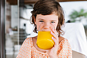 Braunhaariges Mädchen im Vorschulalter schaut in die Kamera und trinkt leckeren Orangensaft aus einem Glas in einer Cafeteria