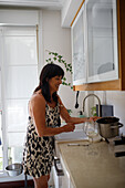 Happy mature brunette in dress smiling and turning on faucet over sink in light kitchen at home