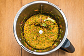 Top view of blender bowl with green fresh mix of chopped vegetables and herbs for preparing healthy appetizer in kitchen