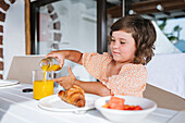 Brown haired child sitting at table and pouring orange juice into glass during breakfast with croissant and fruit salad in cafe